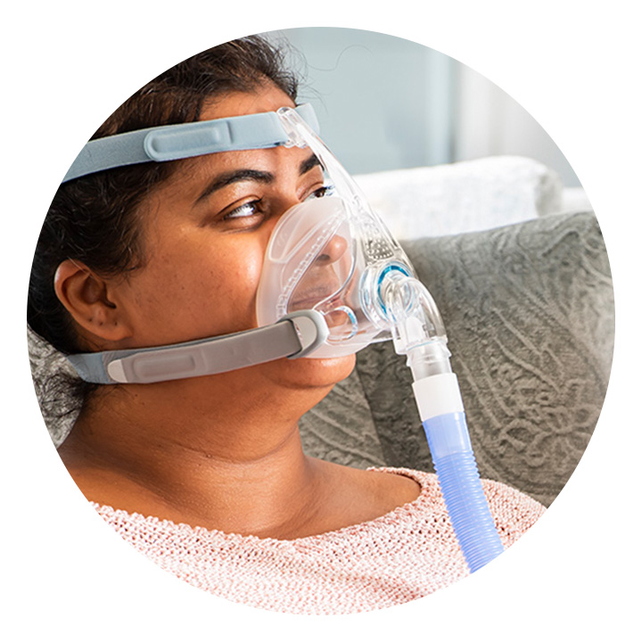 A female patient is sitting in a chair wearing an F&P mask attached to a blue breathing tube